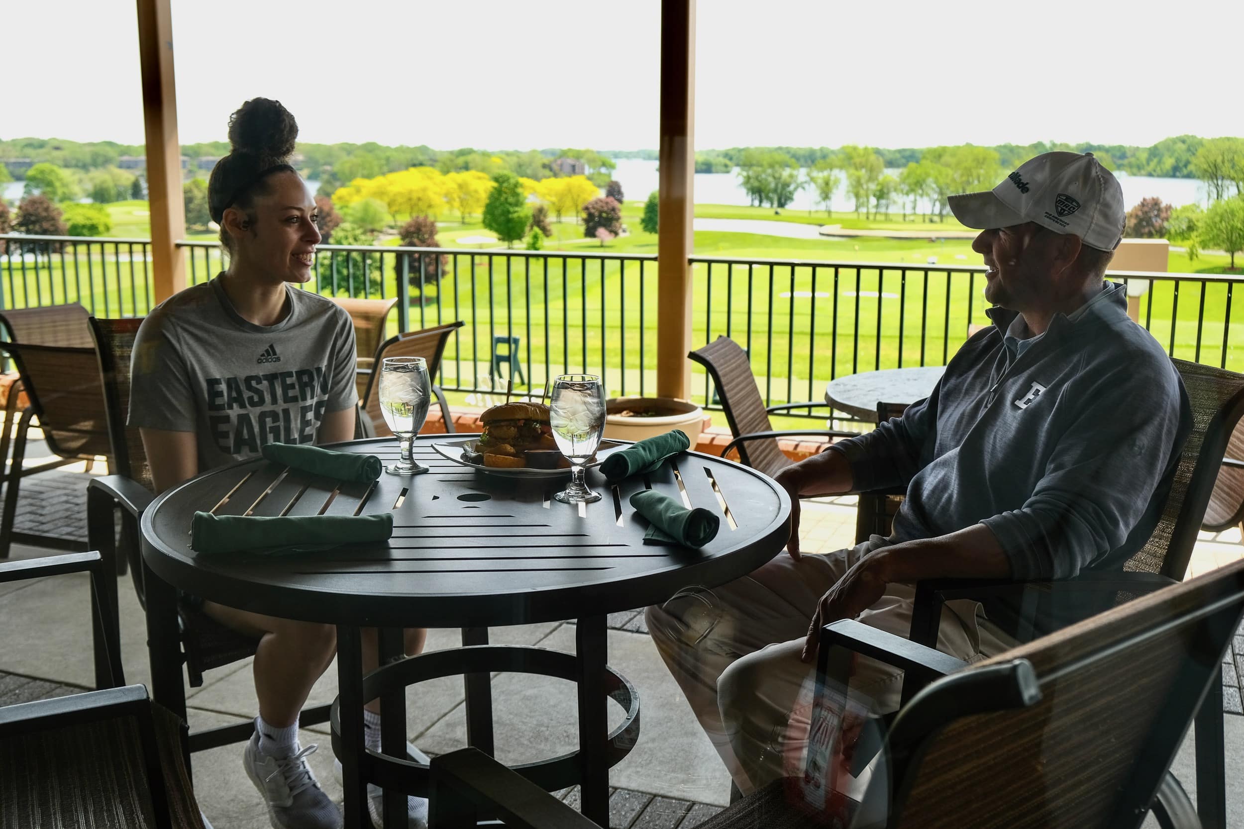 Two people sit at a round patio table with drinks, overlooking a golf course at a scenic golf resort. They are engaged in conversation against the sunny backdrop of Ypsilanti's charming landscape.