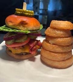 Burger with lettuce, tomato, onion, pickles, and bacon on a bun, next to a stack of onion rings on a white plate.