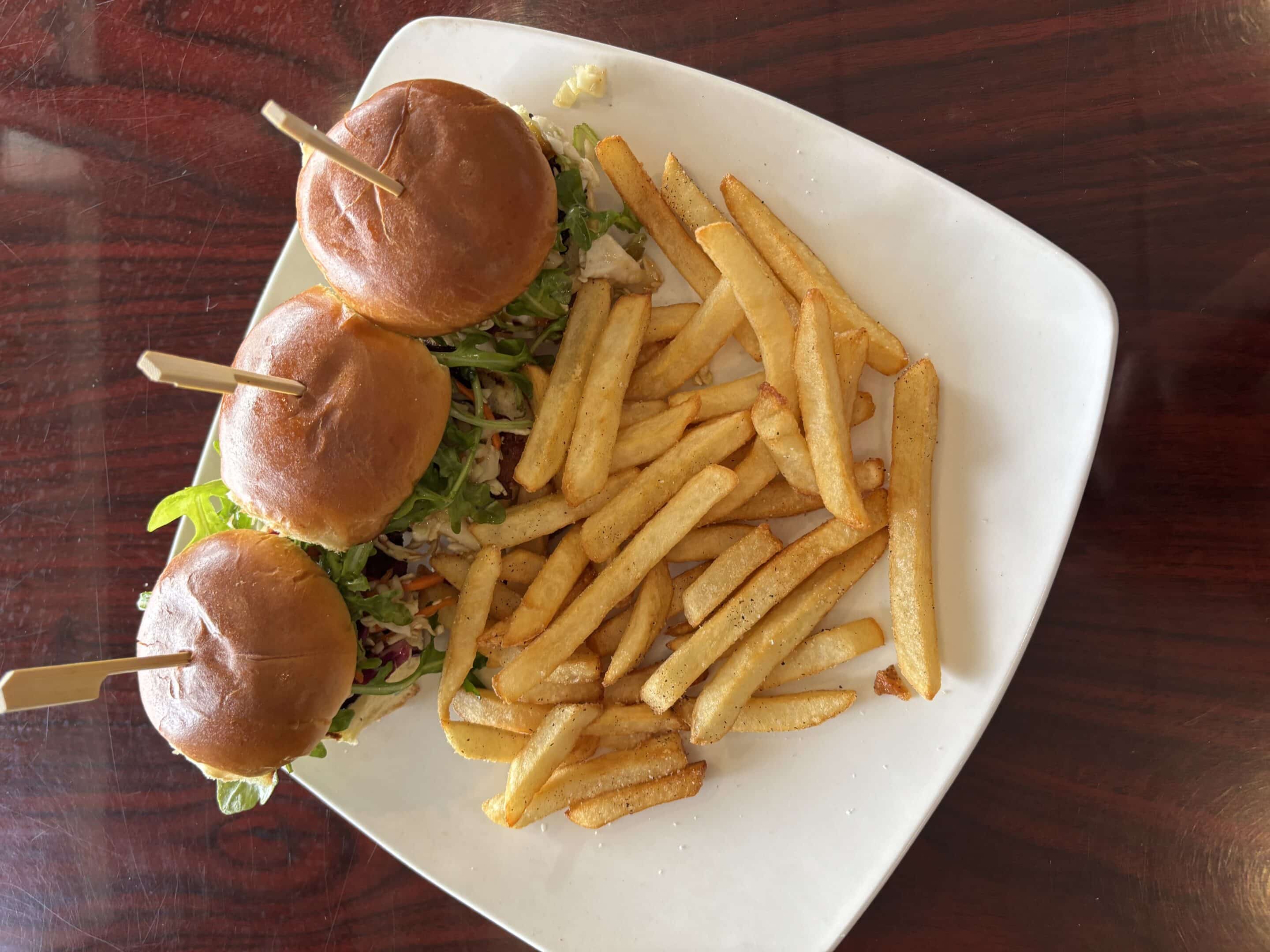 A white plate with three mini burgers on buns, each held with a wooden pick, and a serving of French fries on a wooden table.